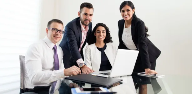 Two business women and two buisiness men dressed in suits stand over a laptop, smiling and looking straight at you.